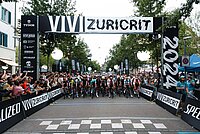 Rennradfahrer stehen mit ihrern Rädern an einer Startlinie. Darüber hängt ein Banner mit der Aufschrift Vivi Zuricrit.