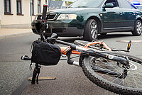 oranges Mountainbike mit lenkertasche liegt vor dunkelgrünem Auto auf der Strasse nach einem Unfall