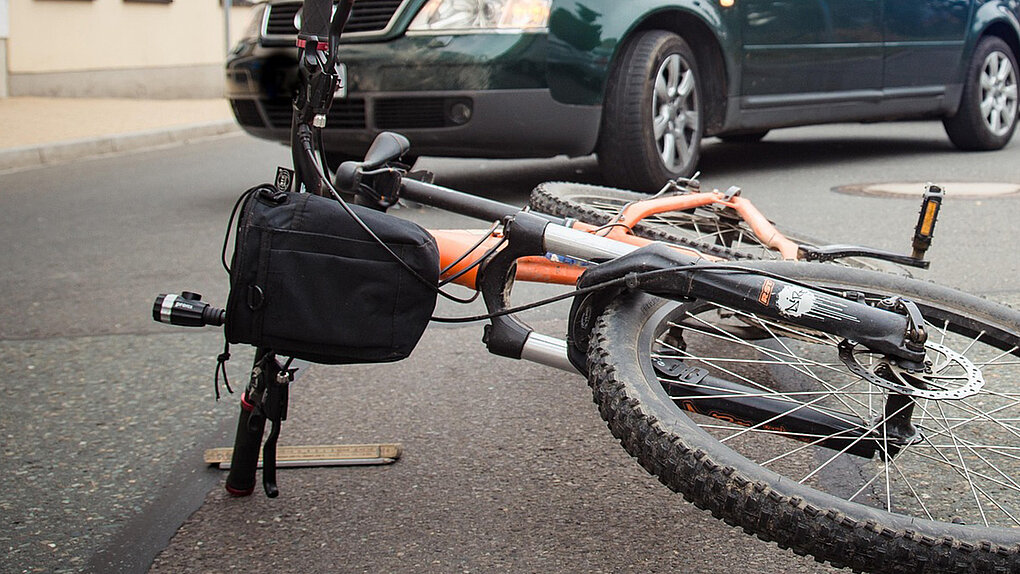 oranges Mountainbike mit lenkertasche liegt vor dunkelgrünem Auto auf der Strasse nach einem Unfall