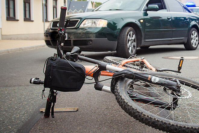 oranges Mountainbike mit lenkertasche liegt vor dunkelgrünem Auto auf der Strasse nach einem Unfall