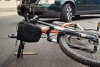 oranges Mountainbike mit lenkertasche liegt vor dunkelgrünem Auto auf der Strasse nach einem Unfall