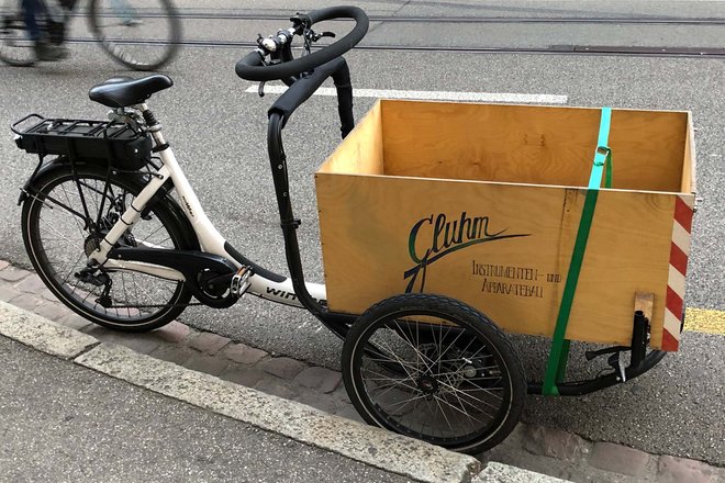 Forschende der Universität Lausanne und der EPFL führen eine Umfrage zur Nutzung von Cargobikes in der Schweiz durch. Dreirädriges Cargobike mit Holzkiste steht auf einer Strasse. 