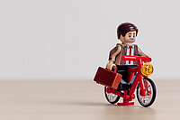 Legomännchen im Anzug mit Aktenkoffer auf Legovelo vor beigem Hintergrund.