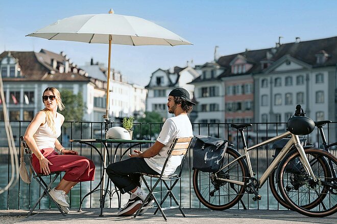 Eine Frau und ein Mann sitzen an einem Bistrotisch am Ufer eines Flusses.
