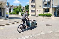 Ein Mann mit Baseballcap fährt auf einem Lastenrad. In der Kiste vorne sitzt ein Kind.