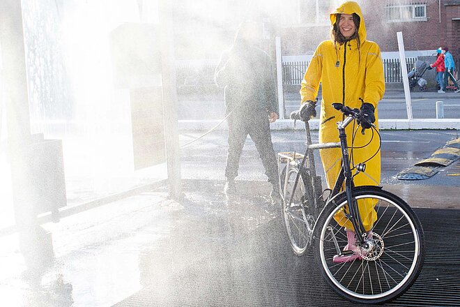 Eine Person steht mit einem gelben Regenoverall und dem Fahrrad unter einer Wasserbrause.