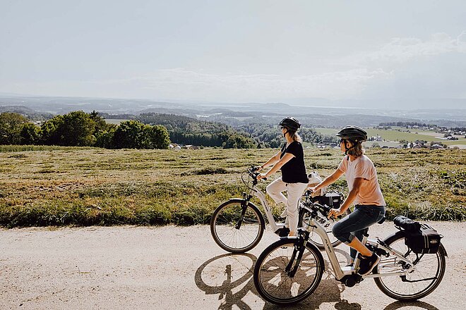 Zwei Frauen fahren auf E-Bikes auf einer Landstrasse. Im Hintergrund grüne Hügel.