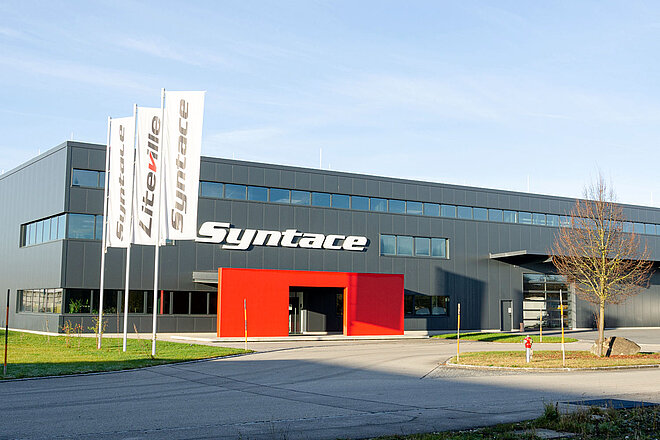 Graue Lagerhalle mit Aufschrift Syntace. Im Vordergrund Flaggen mit der Aufschrift Liteville.