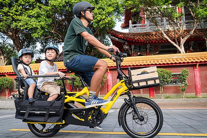 Ein Mann fährt auf einem gelben E-Bike. Auf dem langen Gepäckträger zwei Kinder in Kindersitzen.