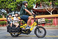 Ein Mann fährt auf einem gelben E-Bike. Auf dem langen Gepäckträger zwei Kinder in Kindersitzen.