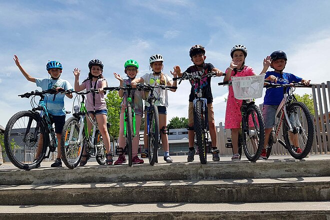 Pro Velo verstärkt bei Bike2School die Digitalisierung. Kinder stehen mit Velos auf einer Treppe und winken.