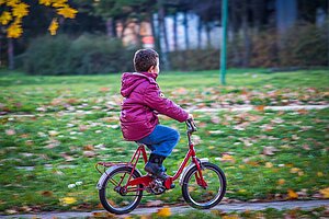 Kind mit pinker Jacke auf rotem Velo fährt einer Wiese entlang.