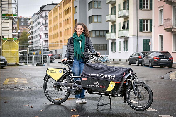Eine Frau mit Schwarzer Jacke steht hinter einem Lastenrad auf der Strasse.