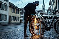 Ein Mann mit Maske und Kapuze schneidet mit einem Winkelschleifer das Schloss eines Fahrrads durch.