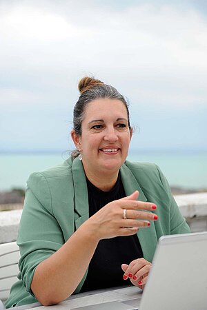 Eine Frau mit grüner Jacke sitzt an einem Tisch. Vor ihr steht ein Laptop.