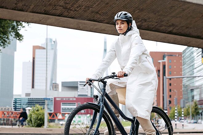 Wer das Fahrrad im Alltag nutzt, möchte funktionelle und praktische Kleider, die auch im Büro oder an der Uni eine gute Figur machen. Funktionelle Kleidung für den Alltag auf zwei Rädern. Eine Frau fährt mit einem weissen Mantel auf dem Fahrrad.