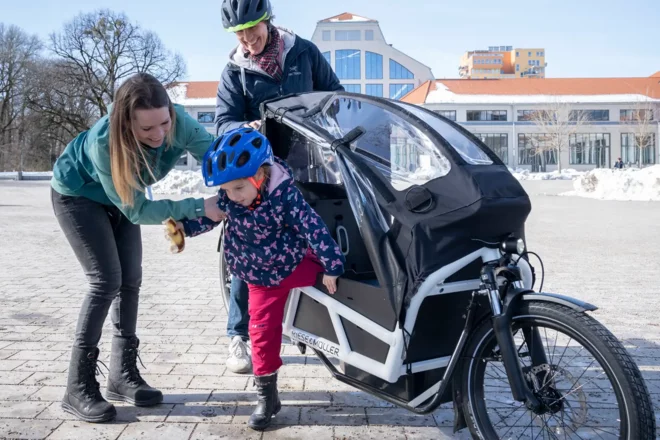 Ein Kind mit Fahrradhelm steigt aus einem Lastenrad aus. Die Eltern stehen daneben. 