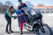 Ein Kind mit Fahrradhelm steigt aus einem Lastenrad aus. Die Eltern stehen daneben. 