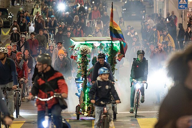 Velofahrende mit bunten Anhängern und bunten Rädern fahren auf einer Strasse.