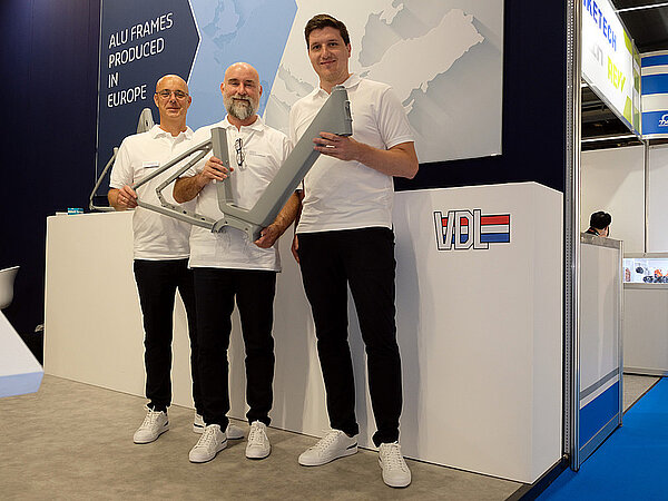 Qwic E-Bike Konkurs. Drei Männer mit weissen T-Shirts halten einen Fahrradrahmen.