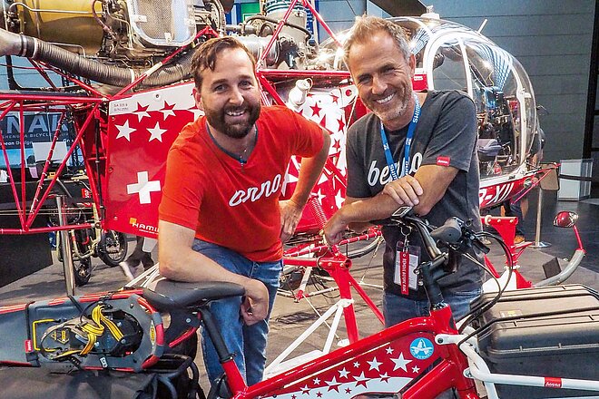 Zwei Männer mit T-Shirts auf denen Benno-Bikes steht stehen vor einem Helikopter der Air Zermatt