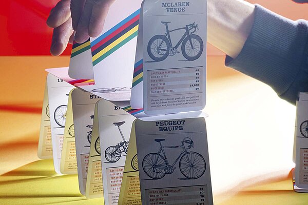 Velofahren ist nicht ganz so günstig, wie gemeinhin angenommen wird. Dafür sind viele Faktoren verantwortlich. Kartenhaus aus Veloquartett vor gelbrotem Hintergrund.