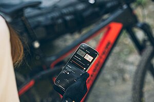 Sram Eeagle Powertrain E-Bike Antrieb. Eine Person mit Bikehandschuhen hält ein Smartphone in der Hand.