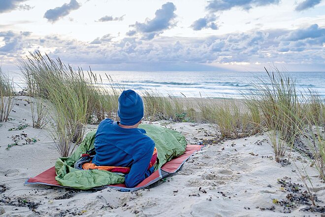 Auf der Tour eine Nacht mit Meeresrauschen verbringen? Der Biwaksack machts möglich. Velojournal hat Biwaksäcke getestet. Ein Mann liegt in einem Biwaksack am Strand.