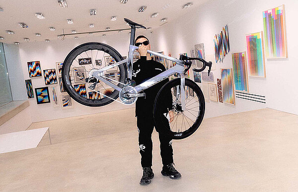 Ein Mann mit schwarzer Kleidung hält ein Rennrad in die Höhe.