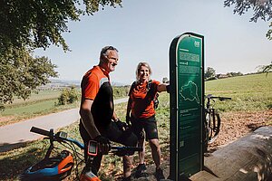 Eine Frau und ein Mann mit Fahrradbekleidung stehen neben einer grünen Infotafel.