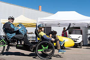 Zwei Personen mit Radhelmen sitzen auf einen Liege-Trike. 