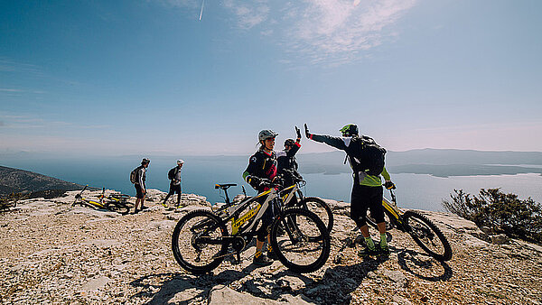 Drei Biker klatschen sich in die Hände. Im Hintergrund ist das Meer zu sehen.