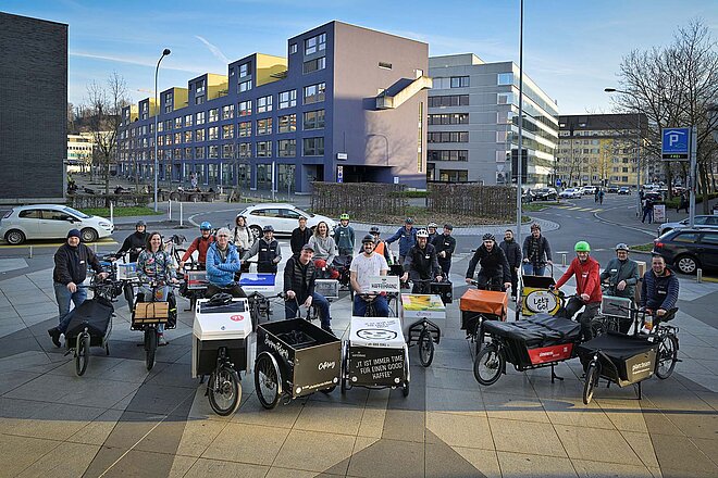 Alber Koechlin Stiftung AKS clever unterwegs. Eine Gruppe von Personen steht mit Cargobikes auf einem Platz.