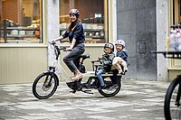 Aloo Diavelo Lontail Cargobikes. Eine Frau fährt auf einem Longtail-Lastenrad mit zwei Kindern in Kindersitzen.