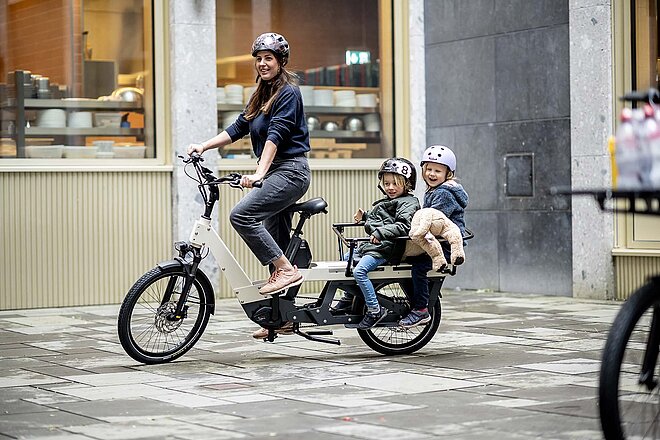 Aloo Diavelo Lontail Cargobikes. Eine Frau fährt auf einem Longtail-Lastenrad mit zwei Kindern in Kindersitzen.