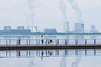 Eine Frau steht auf einem Steg in der Mitte eines Sees. Im Hintergrund rauchen Kamine