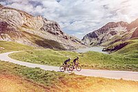 Zwei Rennradfahrer radeln auf einer pitoresken Bergstrass. Im Hinterfrund Berge und ein See