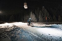 Ein Biker fährt mit dem Bike in der Nacht auf einer beleuchteten Skipiste. 