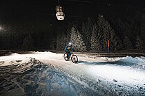 Ein Biker fährt mit dem Bike in der Nacht auf einer beleuchteten Skipiste. 