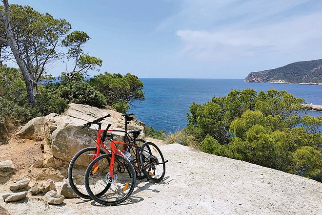 Schöne und ruhige Orte sind mit dem Velo leicht zu erreichen. Radfahren in Mallorca. Zwei Fahrräder stehen an einen Stein angelehnt. Im Hintergrund das Meer.