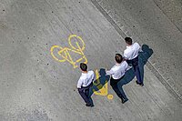 Drei Männer im Anzug laufen auf einem Weg, der mit einen Fussgänger- und einem Velosymbol gekennzeichnet ist.