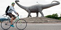 Ein Mann fährt auf einem Velo durch einen Kreisel, auf dem Kreisel steht ein Dinosaurier