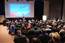 Publikum sitzt in einem Saal und betrachtet eine Präsentation auf einer Leinwand.