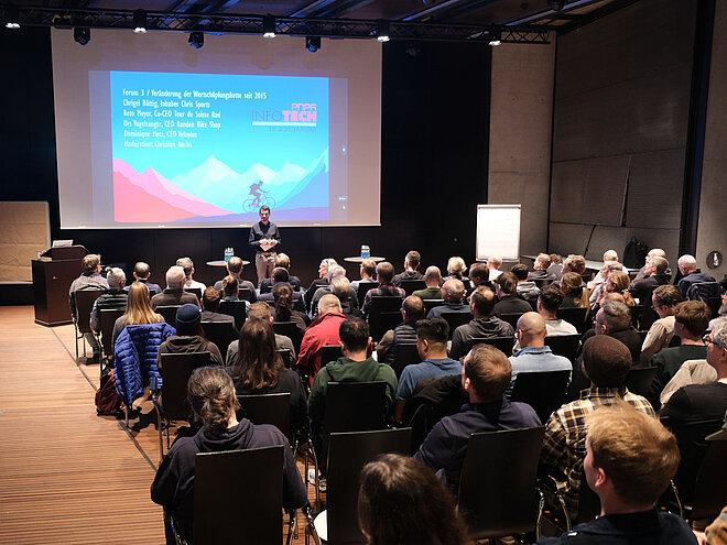 Publikum sitzt in einem Saal und betrachtet eine Präsentation auf einer Leinwand.