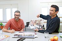 Zwei Männer sitzen an einem Tisch und betrachten Fotos.