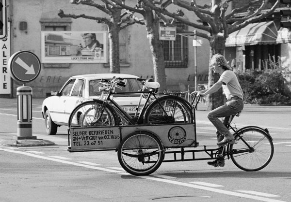 Velociped Cargobike-Center Kriens. Schwarzweiss Foto aus den 80ern, ein Mann fährt auf einem Lastenrad.