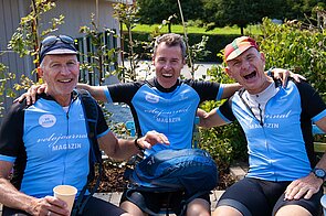 Drei Männer mit Radlerdress sitzen auf einer Bank. 