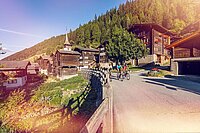 Drei Velofahrer fahren durch Walliser Landschaft an alten Bauernhäusern vorbei. 