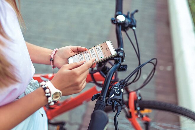 Eine Frau hält ein Smarphone in der Hand und steht neben einem Fahrrad