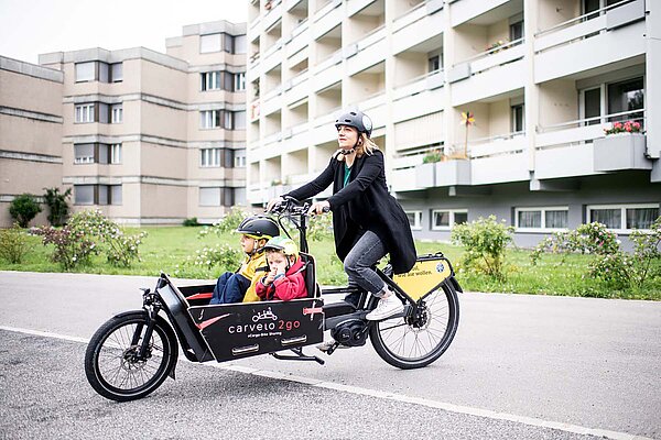 Eine Frau fährt ihre zwei Kinder mit einem Lastenvelo.
